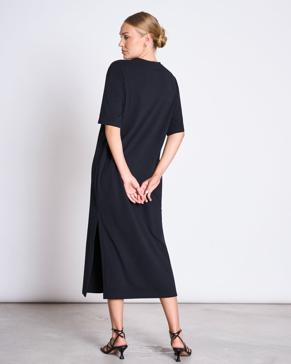 A woman faces away in a jan-n-june SHIRT DRESS CROVIE BLACK, a short-sleeve midi dress with a side slit. She clasps her hands behind her back and wears black strappy heeled sandals against a plain light background.