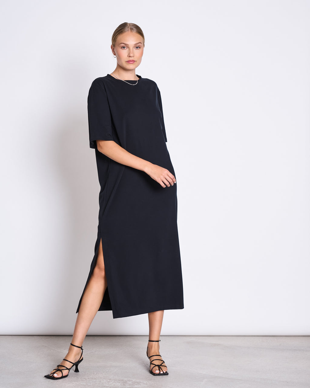 A woman stands against a white wall wearing the jan-n-june SHIRT DRESS CROVIE BLACK, a loose organic cotton midi dress with short sleeves and a side slit, styled with black strappy heels and simple jewelry.