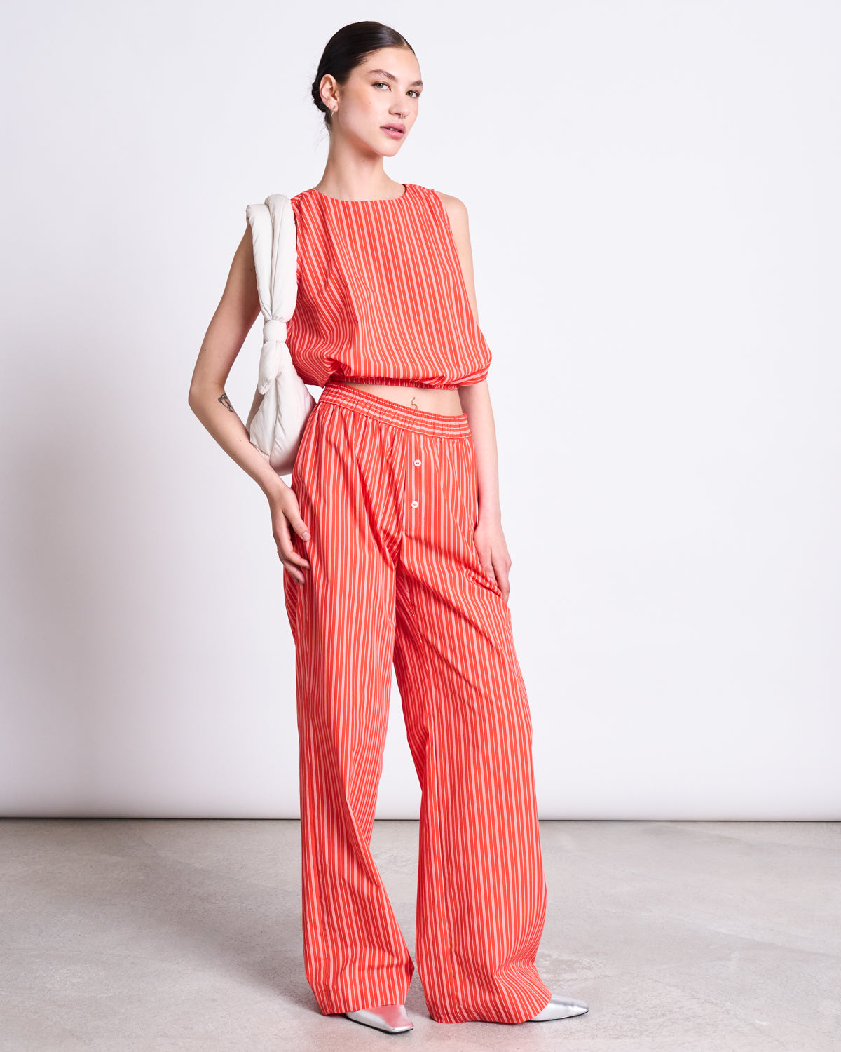A woman poses against a plain backdrop in jan-n-june’s COTTON PANTS SOBY CHERRY RED WHITE STRIPED GOTS, paired with a matching sleeveless top, white shoulder bag, pointed shoes, and her hair tied back.