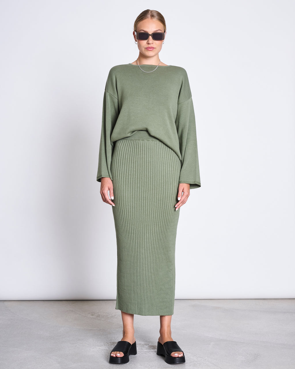 A person in sunglasses wears a sage green long-sleeve sweater with the jan-n-june MIDI KNIT SKIRT EBBA SEA SPRAY GOTS, pairing the look with black platform sandals and a silver necklace on a light gray floor.