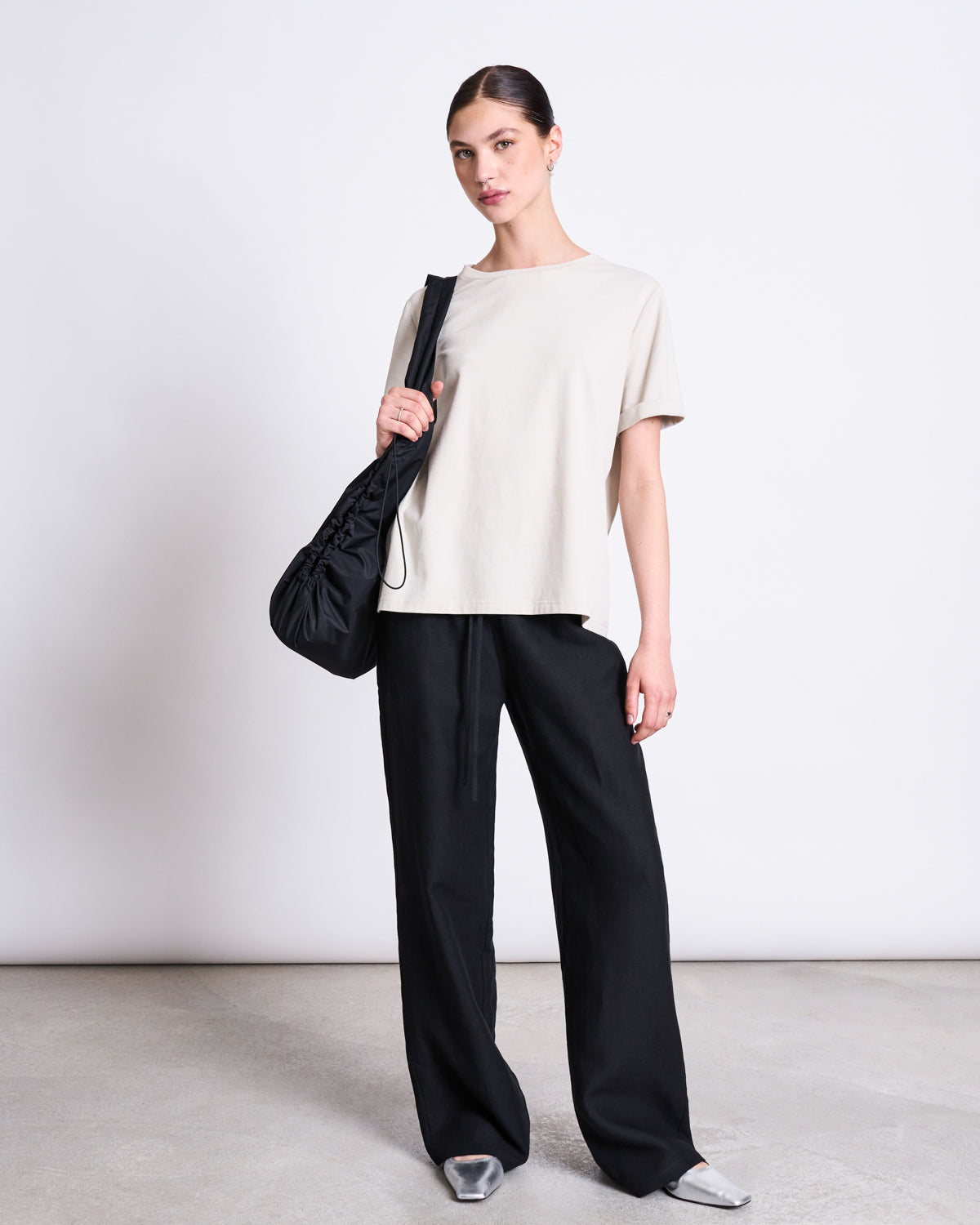 A woman poses against a white background wearing a loose beige tee, wide-leg black pants, silver shoes, and carries the jan-n-june DRAWSTRING SHOPPER LYNE BLACK. Her hair is styled in a neat bun for a chic, modern look.