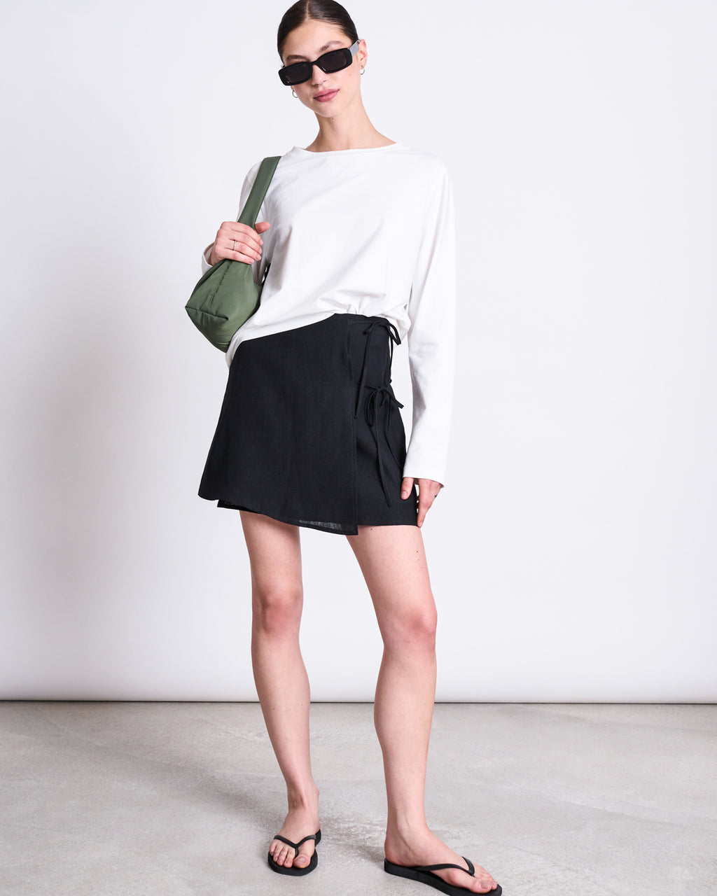A woman stands against a plain background wearing black sunglasses, a white long-sleeve top, the jan-n-june LINEN TIE SKIRT KYS BLACK—a mini linen wrap skirt with side ties—black flip-flops, and a small green shoulder bag.