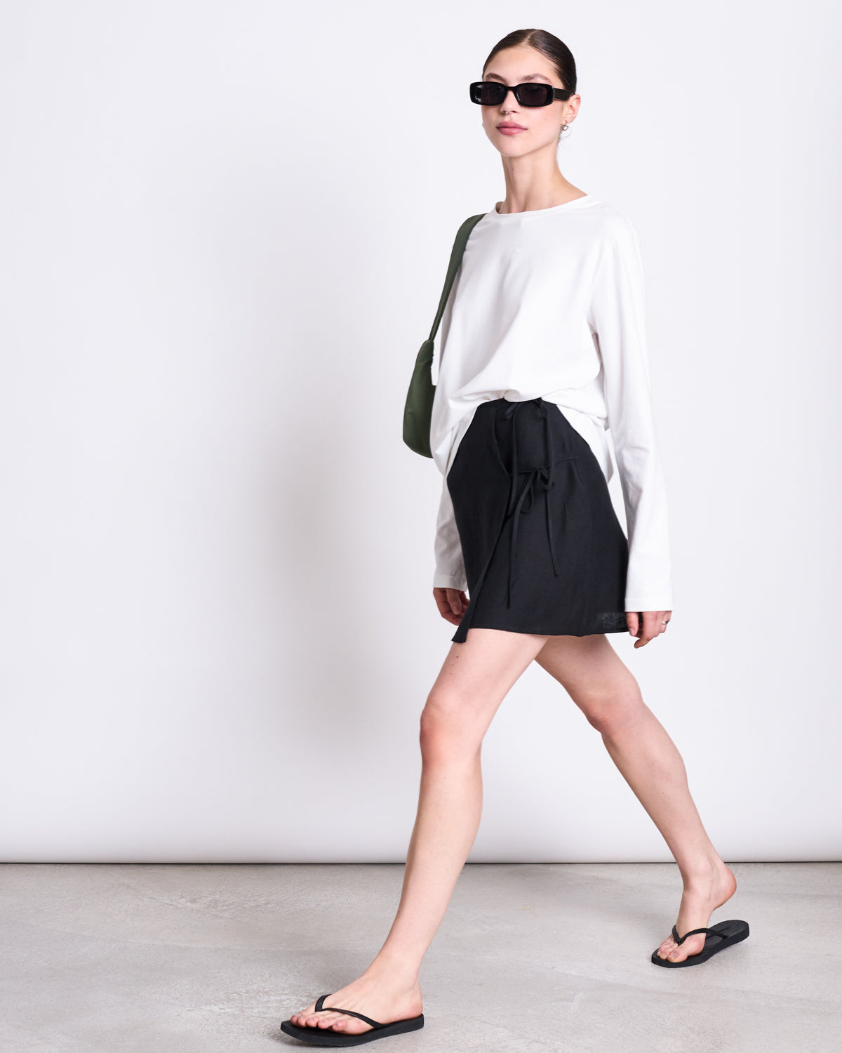 A woman walks confidently against a plain light background wearing the jan-n-june BRUSHED LONGSLEEVE BOY WHITE GOTS shirt, a black skirt, flip-flops, sunglasses, and carrying a green shoulder bag.