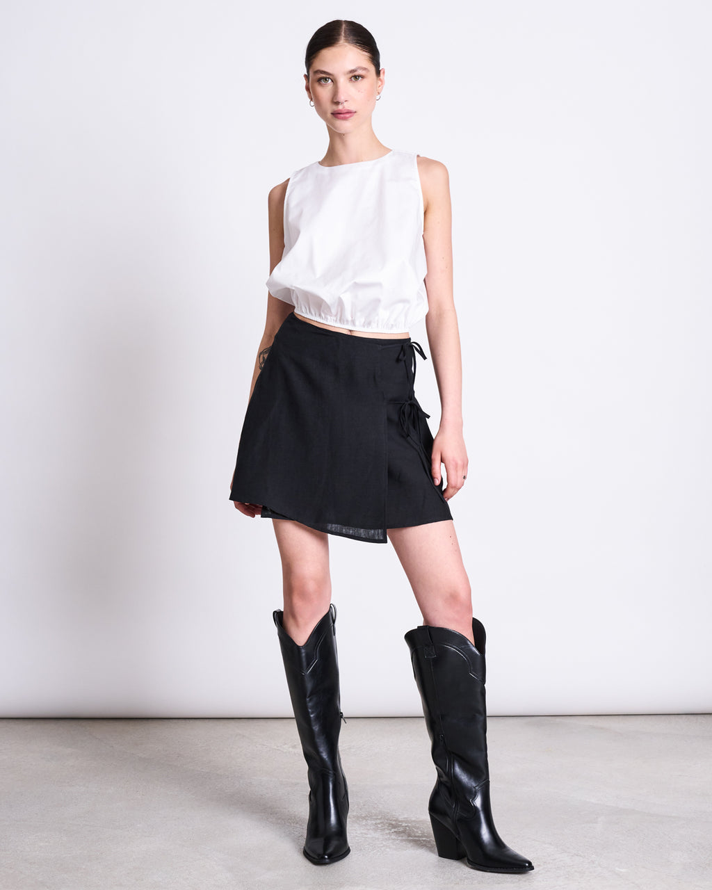 A woman poses in a studio against a white background, wearing the jan-n-june LINEN TIE SKIRT KYS BLACK—a mini-length black linen wrap skirt—paired with a sleeveless white top and tall black cowboy boots. Her hair is tied back.