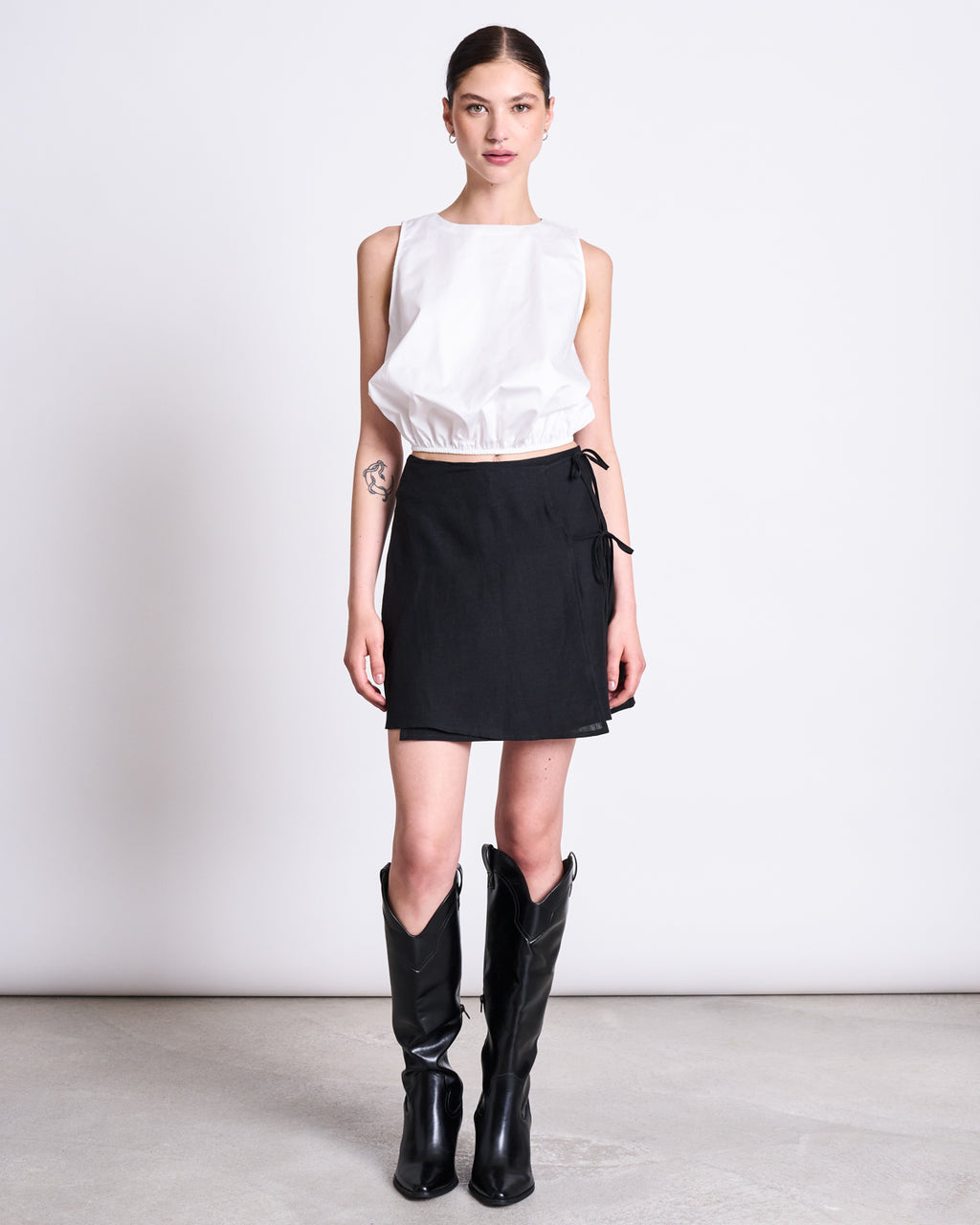 A woman stands against a white wall wearing the jan-n-june LINEN TIE SKIRT KYS BLACK, a mini European linen skirt with side ties, paired with a sleeveless top and knee-high black boots. Her hair is tied back, showing a small left forearm tattoo.
