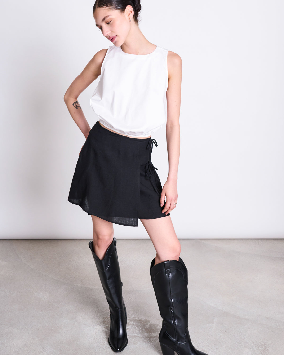 A woman poses against a plain backdrop in a sleeveless white top, knee-high black boots, and the jan-n-june LINEN TIE SKIRT KYS BLACK—a mini-length wrap skirt crafted from European linen. She stands with one leg forward and her head tilted to the side.