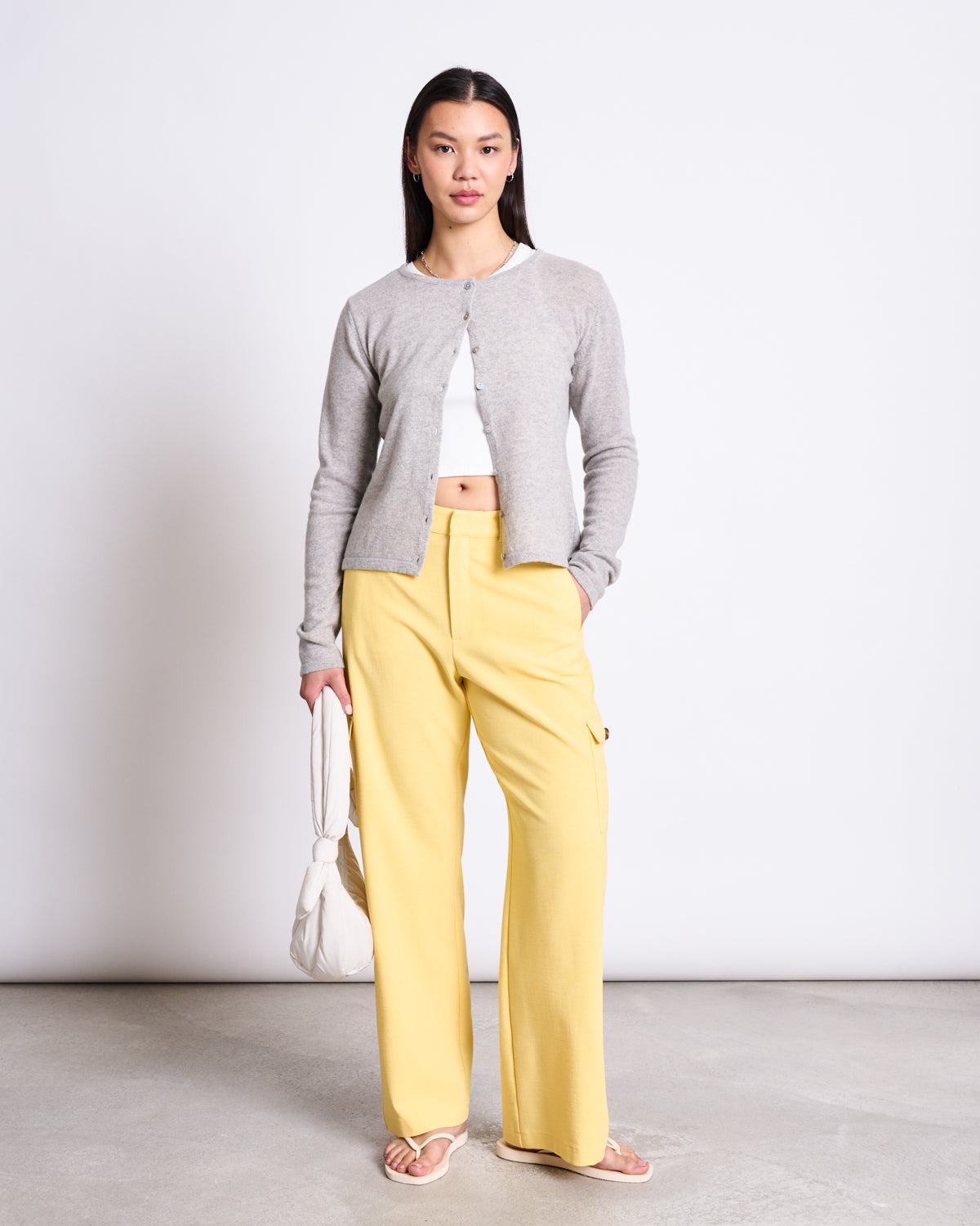 A woman with straight dark hair and a neutral expression stands against a white wall, wearing jan-n-june CARGO PANTS FANO BUTTERCUP, a light gray cardigan over a white crop top, sandals, and holding a white bag.