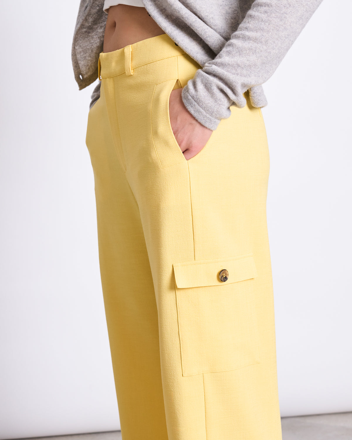 A person wears jan-n-junes CARGO PANTS FANO BUTTERCUP, light yellow wide-leg cargo pants made from recycled polyester, with a side pocket, and a grey long-sleeve top, standing hand-in-pocket against a plain background.