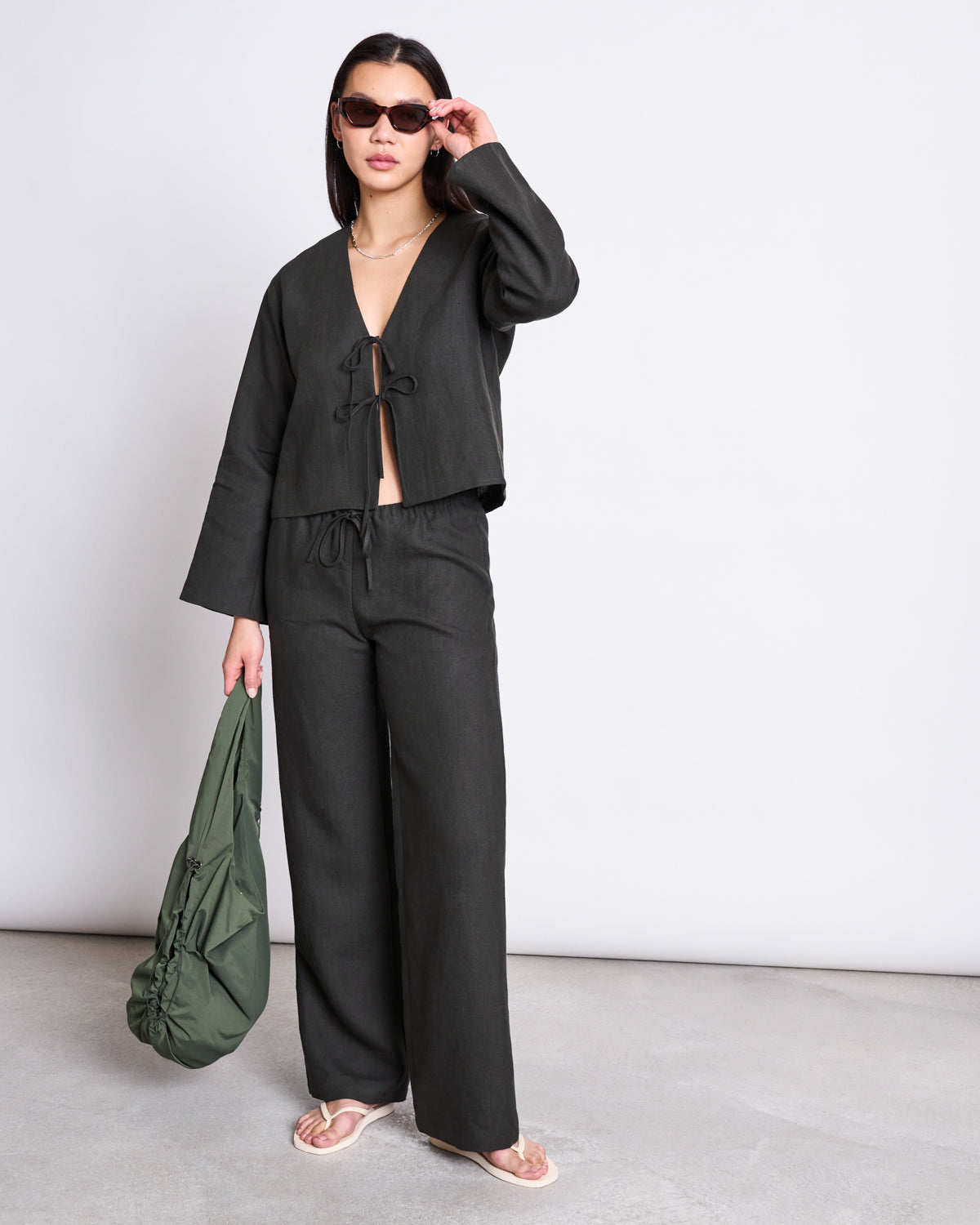 A woman poses against a plain background in the jan-n-june LINEN TIE BLOUSE SKAGEN DARK OLIVE, paired with loose pants, sunglasses, flat sandals, and carrying a large green bag.