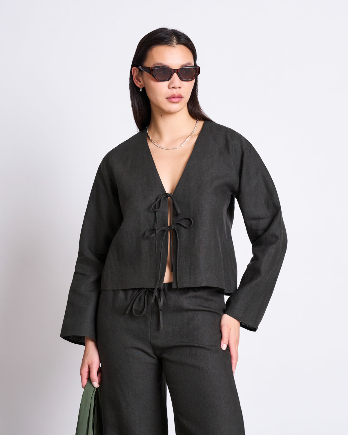 A woman in dark sunglasses and the jan-n-june LINEN TIE BLOUSE SKAGEN DARK OLIVE with matching pants poses confidently against a plain white background.