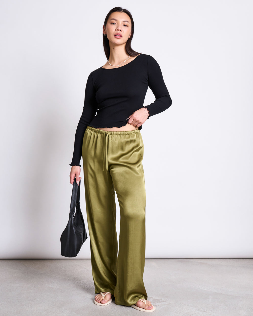 A woman stands against a plain background wearing jan-n-june’s LOOSE SILKY PANTS FRIS WOODY OLIVE, styled with a fitted black long-sleeve top, a black bag, and beige sandals.