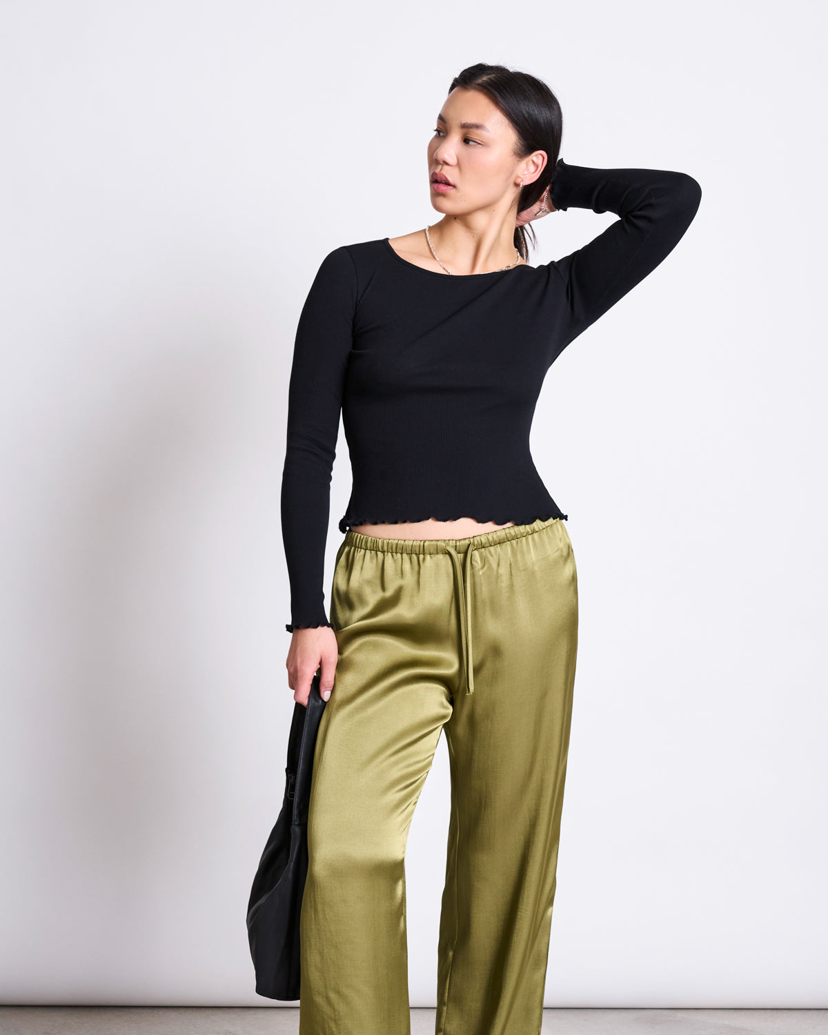 A woman stands against a plain white wall wearing the jan-n-june MINIRIB LONGSLEEVE AVERSA BLACK GOTS, paired with shiny olive green satin pants. She holds a black bag, one hand resting on the back of her head.