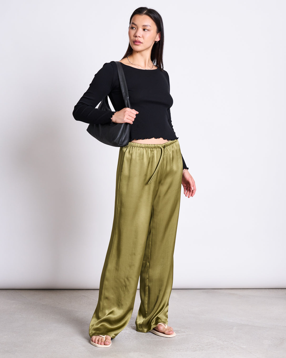 A woman stands indoors wearing jan-n-june LOOSE SILKY PANTS FRIS WOODY OLIVE, a black long-sleeve top, beige sandals, and a black shoulder bag. She looks to the side with a neutral expression.