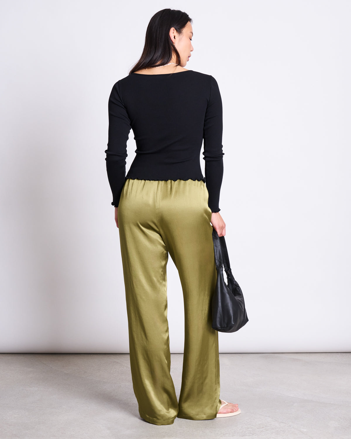 A woman faces away, wearing jan-n-june LOOSE SILKY PANTS FRIS WOODY OLIVE—loose, olive-green vegan silk pants—paired with a black fitted top. She holds a black handbag, standing on a light gray floor against a plain white wall.