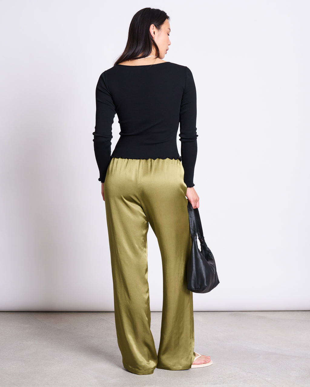 A woman with long dark hair stands facing away against a plain background, wearing the jan-n-june MINIRIB LONGSLEEVE AVERSA BLACK GOTS top and holding a black bag in her left hand, paired with loose olive green satin pants.
