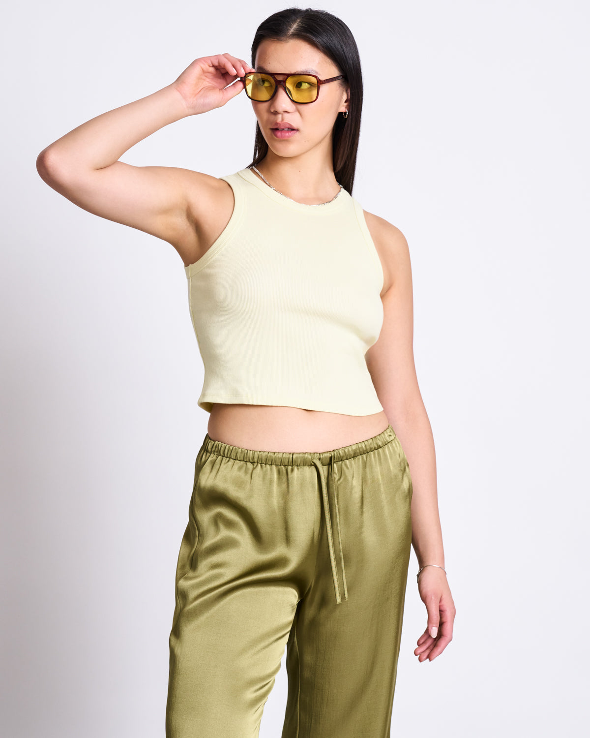 A woman stands against a white background, looking to the side and adjusting her yellow sunglasses while wearing jan-n-junes LOOSE SILKY PANTS FRIS WOODY OLIVE with a sleeveless cream crop top.