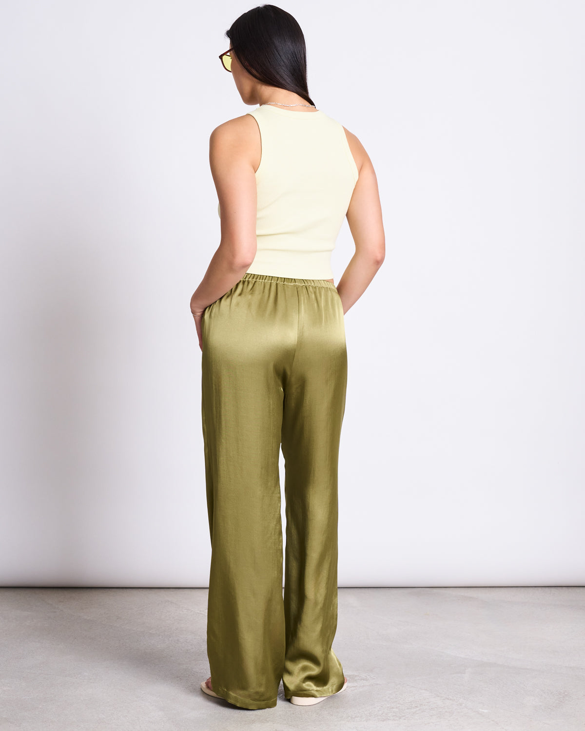 A woman with long dark hair wears jan-n-junes LOOSE SILKY PANTS FRIS WOODY OLIVE—loose, olive vegan silk low-waist pants—with a sleeveless cream top, sunglasses, and sandals, standing against a plain white background.