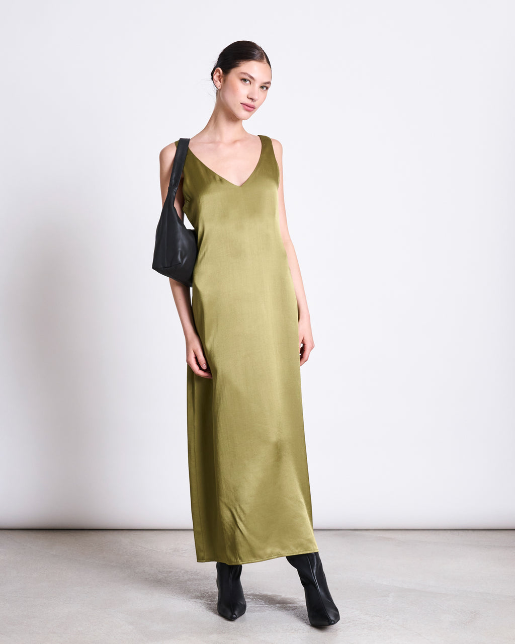 A woman poses against a plain background in the jan-n-june MAXI DRESS BENVIE WOODY OLIVE, styled with black ankle boots and a matching shoulder bag. Her hair is pulled back as she gazes at the camera.