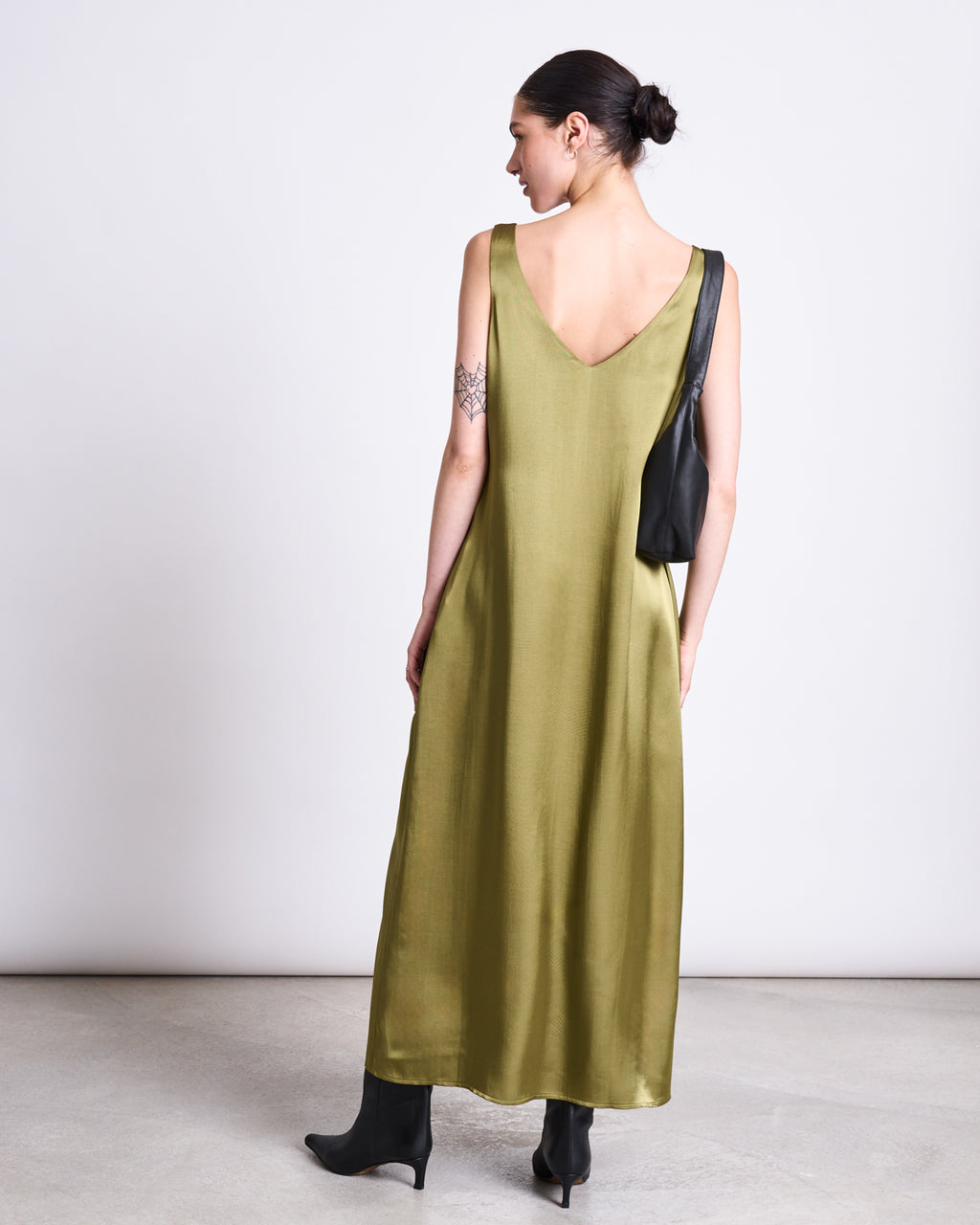 A woman with dark hair in a bun wears the jan-n-june MAXI DRESS BENVIE WOODY OLIVE, featuring a deep V-back. She pairs it with black boots and a black handbag, posing against a light background.