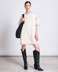 A woman with tied-back hair poses against a plain background in the jan-n-june T-SHIRT DRESS HOPLA ECRU GOTS—an oversized organic cotton t-shirt dress—styled with black knee-high cowboy boots and a black shoulder bag.