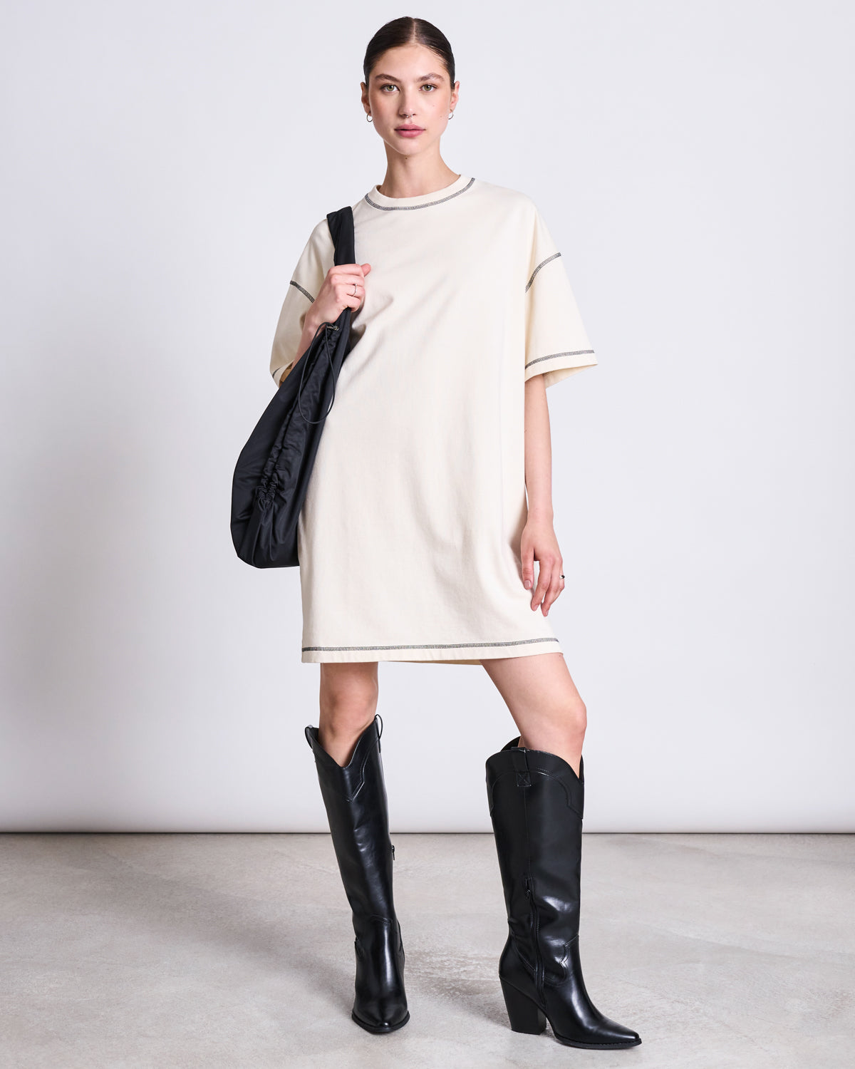 A woman with tied-back hair poses against a plain background in the jan-n-june T-SHIRT DRESS HOPLA ECRU GOTS—an oversized organic cotton t-shirt dress—styled with black knee-high cowboy boots and a black shoulder bag.