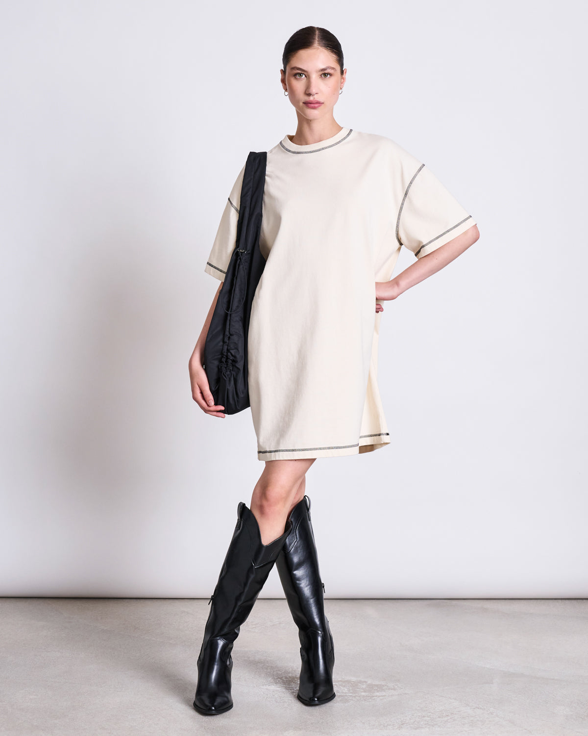 A woman poses against a plain background wearing the jan-n-june T-SHIRT DRESS HOPLA ECRU GOTS. She pairs the oversized cream dress with black knee-high boots and a black shoulder bag, her hair pulled back and one hand on her hip.
