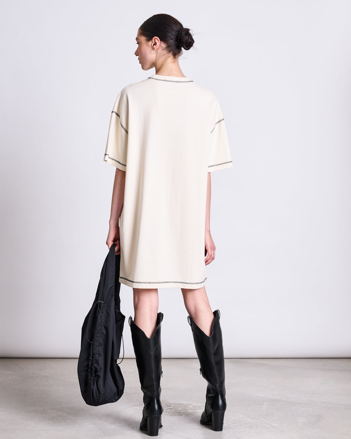 A person stands facing away, wearing the jan-n-june T-SHIRT DRESS HOPLA ECRU GOTS—an oversized beige organic cotton dress—with black knee-high boots and a black bag, hair in a bun, set against a plain light background.
