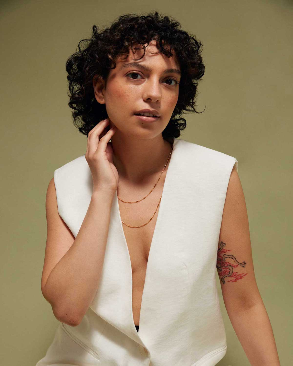 A person with short, curly dark hair and a dragon tattoo on their upper arm sits against a plain background, wearing a sleeveless white vest and sustainable jewelry: the jan-n-june DEAR DARLING BERLIN DELICATE NECKLACE WITH FINE BARS GOLD.