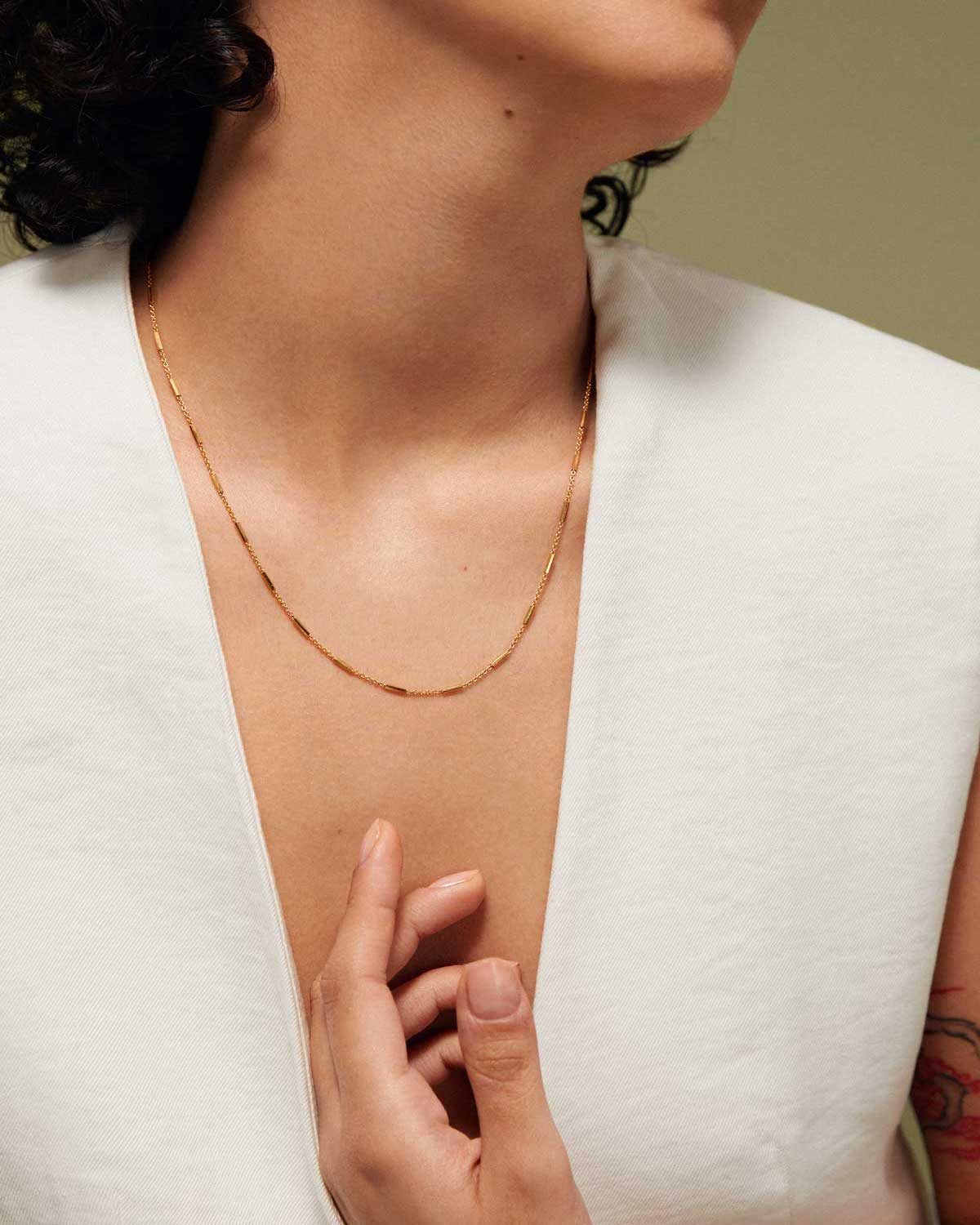 A woman in a white sleeveless top wears the jan-n-june DEAR DARLING BERLIN DELICATE NECKLACE WITH FINE BARS GOLD, gently touching her collarbone. Only her neck, lower face, and hand are visible for a minimalist, elegant style.
