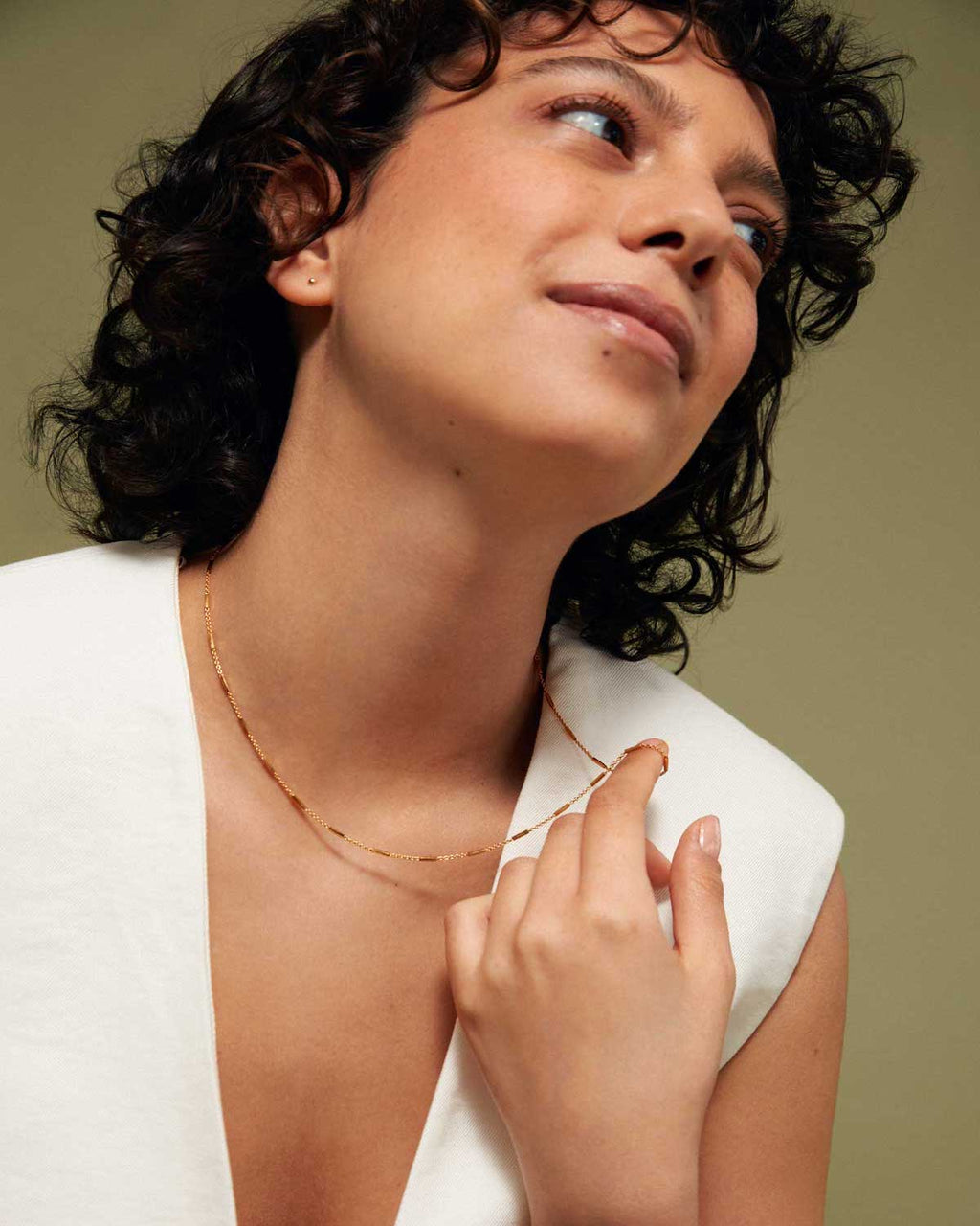 A person with short, curly dark hair in a white sleeveless top smiles softly and looks upward, holding the jan-n-june DEAR DARLING BERLIN DELICATE NECKLACE WITH FINE BARS GOLD against their chest. The background is muted olive.