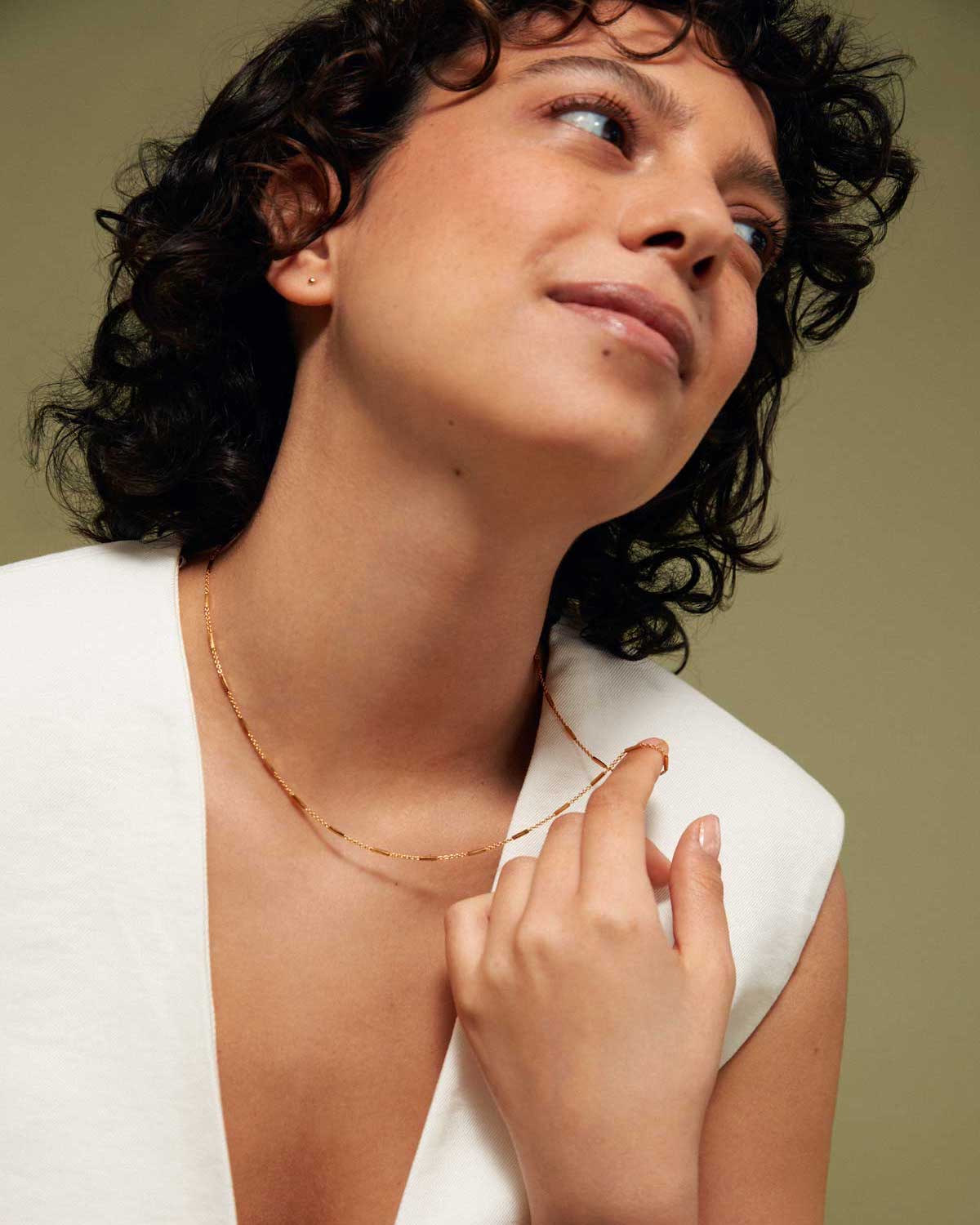 A person with short, curly dark hair in a white sleeveless top smiles softly and looks upward, holding the jan-n-june DEAR DARLING BERLIN DELICATE NECKLACE WITH FINE BARS GOLD against their chest. The background is muted olive.