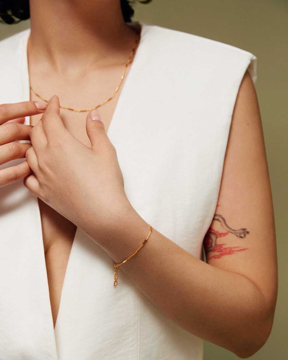 A person wearing a sleeveless white top adjusts the jan-n-june DEAR DARLING BERLIN DELICATE NECKLACE WITH FINE BARS GOLD. They also wear a matching gold bracelet, with a red and black tattoo visible on their upper arm against a muted green background.