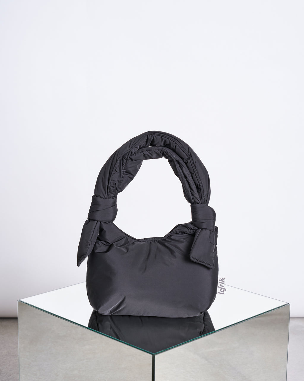 The BAG BIWA PUFFY MINI BLACK by jan-n-june, a black padded handbag with a knotted handle made from recycled fabric, is displayed on a mirrored surface against a plain white background.