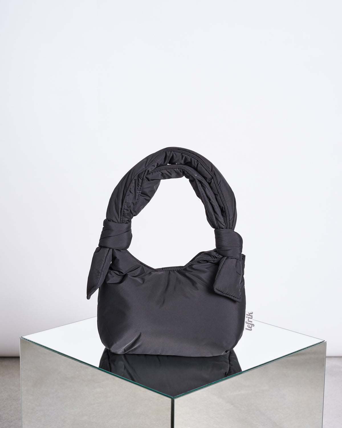 The BAG BIWA PUFFY MINI BLACK by jan-n-june, a black padded handbag with a knotted handle made from recycled fabric, is displayed on a mirrored surface against a plain white background.
