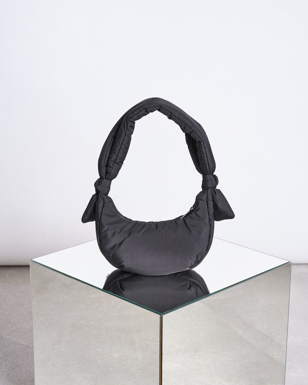 The jan-n-june BAG BIWA PUFFY MICRO BLACK, a crescent-shaped handbag with knotted handles made from recycled polyester, is displayed on a mirrored cube against a plain white background.