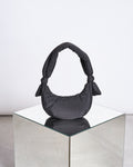 The jan-n-june BAG BIWA PUFFY MICRO BLACK, a crescent-shaped handbag with knotted handles made from recycled polyester, is displayed on a mirrored cube against a plain white background.