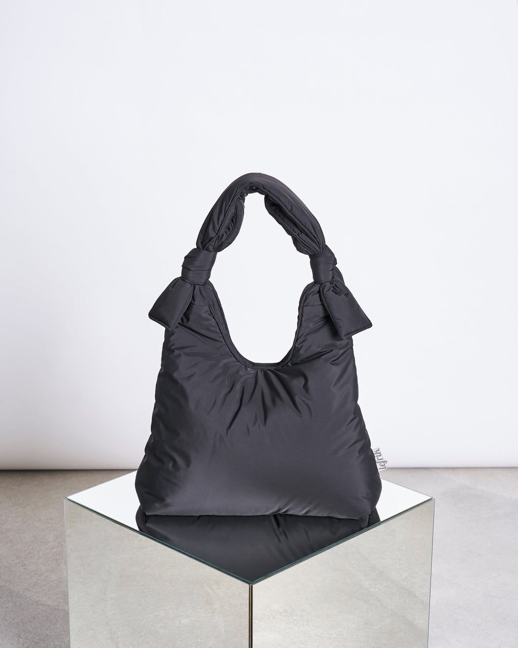 The BAG BIWA PUFFY REGULAR BLACK by jan-n-june, made from recycled plastic fabric with adjustable sliding knot handles, rests on a mirrored cube against a white and gray backdrop.