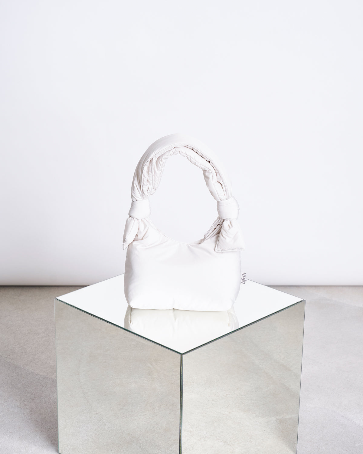 The jan-n-june BAG BIWA PUFFY MINI ICE, a white bag with a soft texture and knotted handle made from recycled polyester, is displayed on a mirrored cube against a plain, light-toned background.