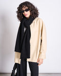 A person with curly hair, sunglasses, a beige button-up jacket, jan-n-june’s WOOLY KNIT SCARF AVAK BLACK in a wool-mohair blend, and black pants stands against a plain white background.