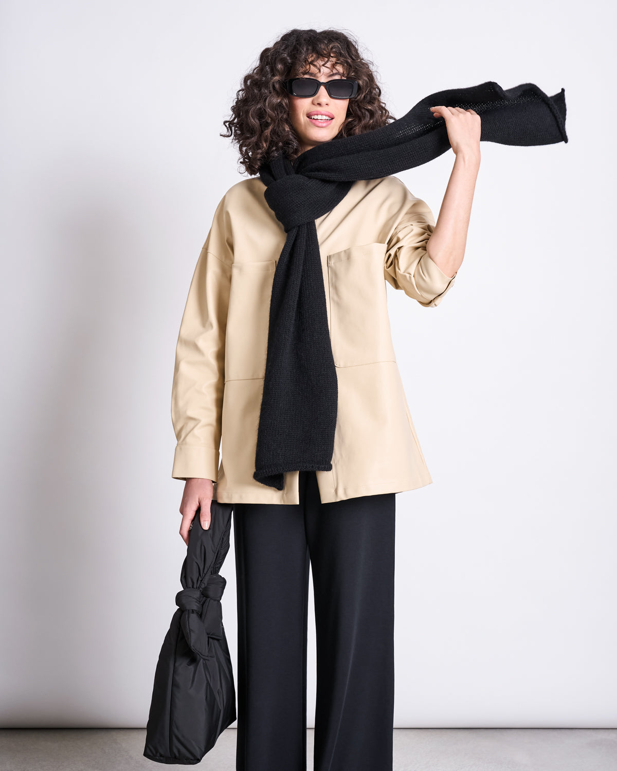 A woman with curly hair in sunglasses, a beige jacket, black wide-leg pants, and a jan-n-june WOOLY KNIT SCARF AVAK BLACK (wool-mohair mix) smiles while holding a black bag. The chic scarf is stylishly draped over her shoulder.