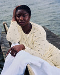 A woman in the jan-n-june POINTELLE PYJAMA CREWNECK BUTTERBOW GOTS, a light yellow organic cotton set with small prints, sits thoughtfully on a wooden dock by the water, resting her head on one hand with a white cloth nearby.