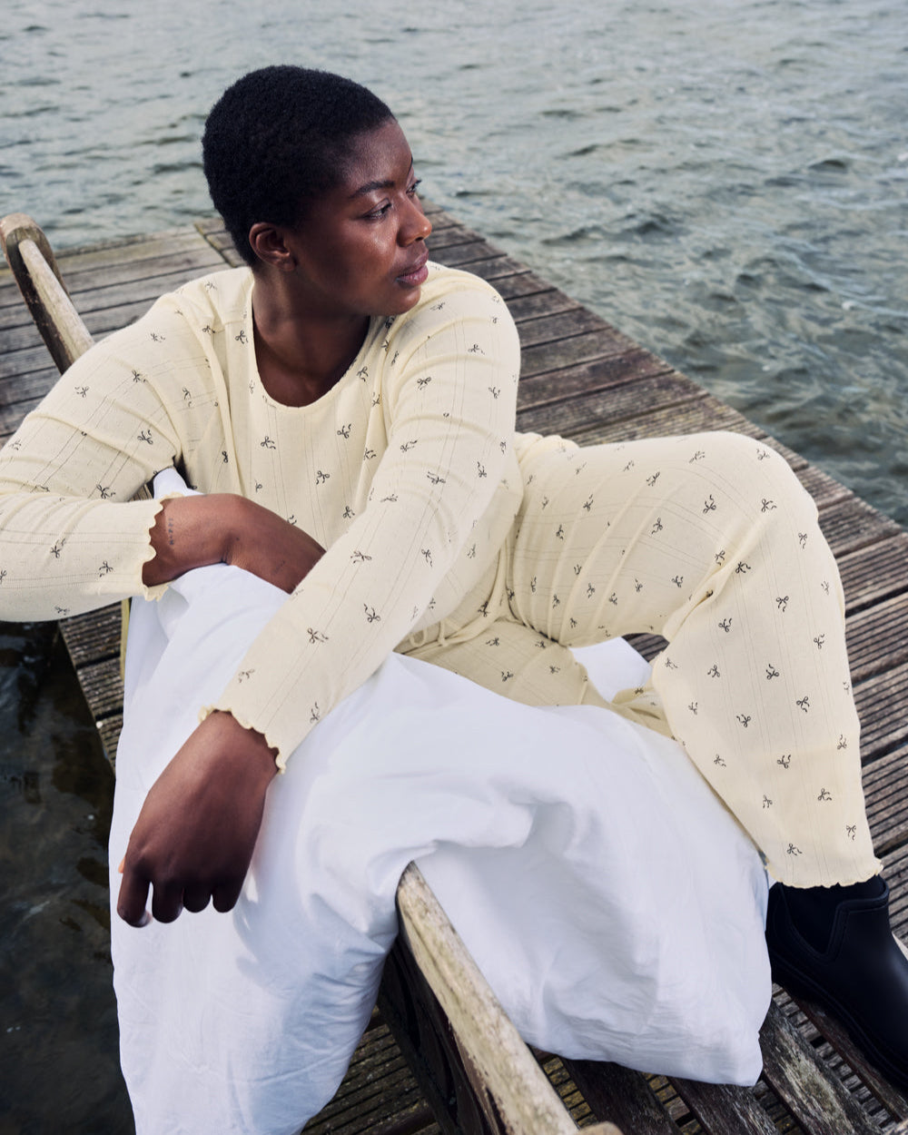 A person in the jan-n-june POINTELLE PYJAMA CREWNECK BUTTERBOW GOTS sits on a wooden dock by the water, holding a white blanket and gazing thoughtfully to the side.