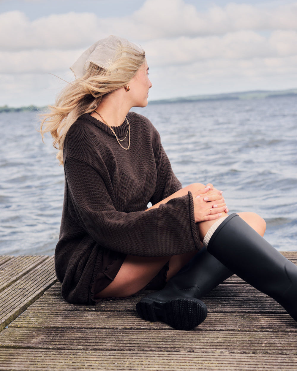 A woman sits on a dock by the water, wearing the jan-n-june JUMPER SONA CHOCOLATE GOTS and black rain boots. A light scarf covers her head as her blonde hair catches the breeze under a cloudy sky.