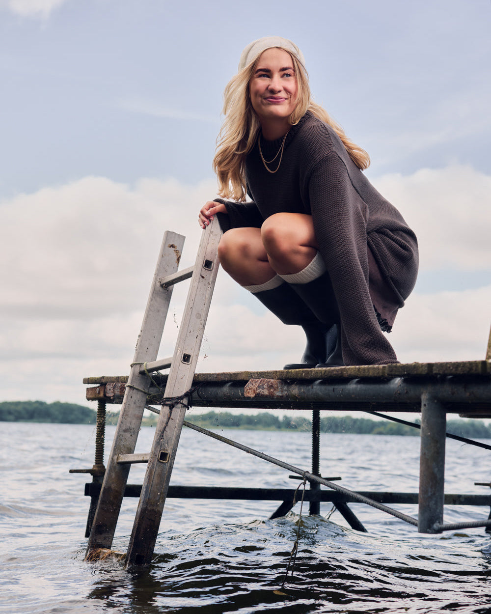 A woman with long blonde hair smiles as she squats on a wooden dock above the water. She wears the jan-n-june JUMPER SONA CHOCOLATE GOTS, along with knee-high socks and boots, under a partly cloudy sky.