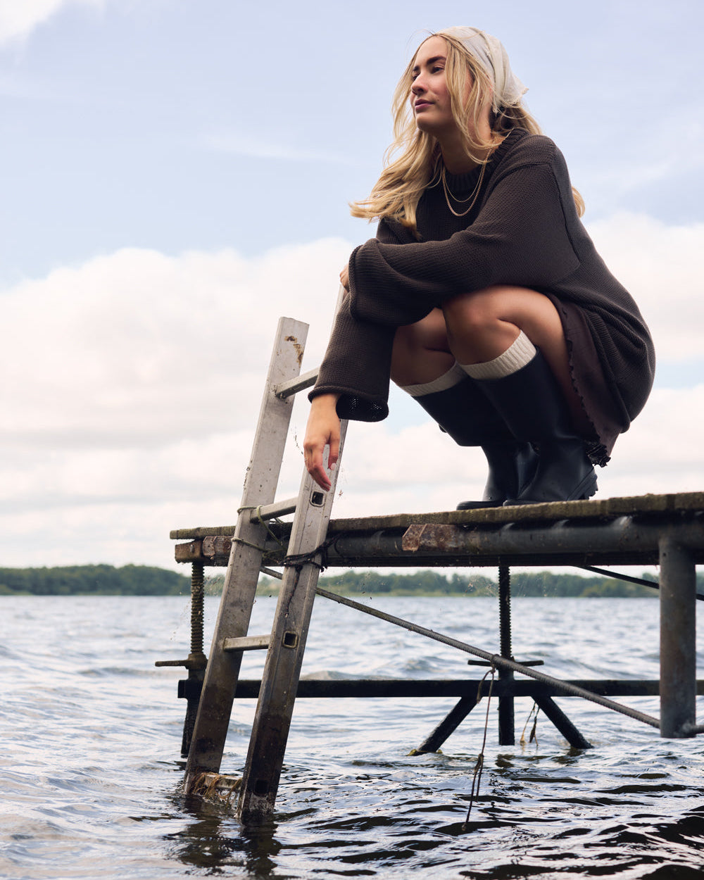 A blonde woman squats on a wooden dock above a lake, holding a metal ladder. She wears the JUMPER SONA CHOCOLATE GOTS by jan-n-june and black boots, with trees and a cloudy sky in the background.