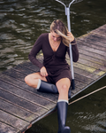 A woman in a brown outfit and dark rain boots, wearing the jan-n-june POINTELLE PYJAMA V-NECK LONGSLEEVE CHOCOLATE GOTS, sits on a wooden dock by the water with a fishing net, her hair partially covering her face.