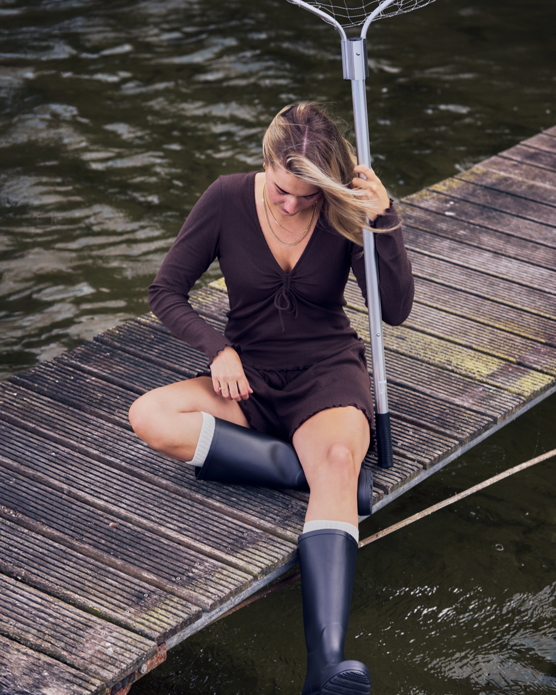 A woman in a brown outfit and dark rain boots, wearing the jan-n-june POINTELLE PYJAMA V-NECK LONGSLEEVE CHOCOLATE GOTS, sits on a wooden dock by the water with a fishing net, her hair partially covering her face.
