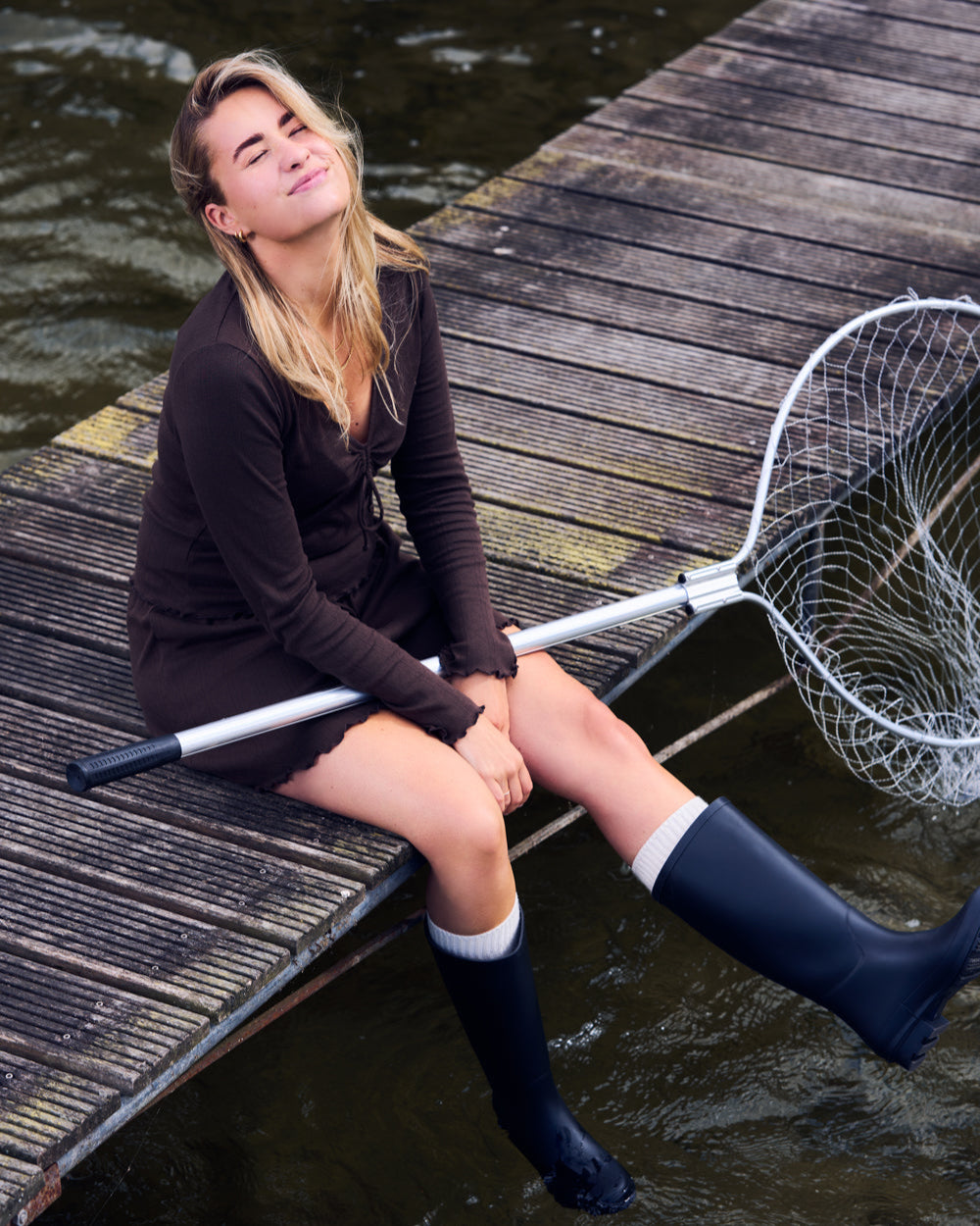 A woman wearing the jan-n-june POINTELLE PYJAMA V-NECK LONGSLEEVE CHOCOLATE GOTS sits on a wooden dock by the water, holding a large fishing net and smiling with her eyes closed.
