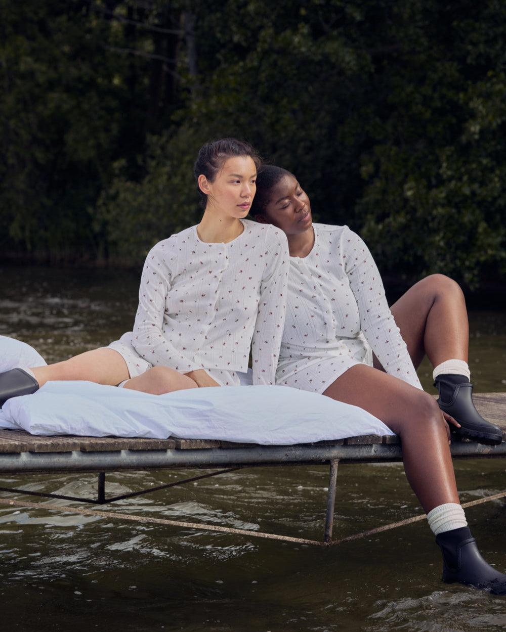 Two women in matching jan-n-june POINTELLE PYJAMA BUTTONED LONGSLEEVE BLOOMY GOTS sets made from certified organic cotton sit on a bed floating near woods, relaxing outdoors as one rests her head on the others shoulder.