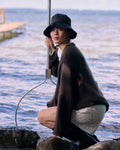 A woman in the jan-n-june JUMPER SONA CHOCOLATE GOTS and a black bucket hat kneels by a serene lakeside dock, holding a large fishing net.